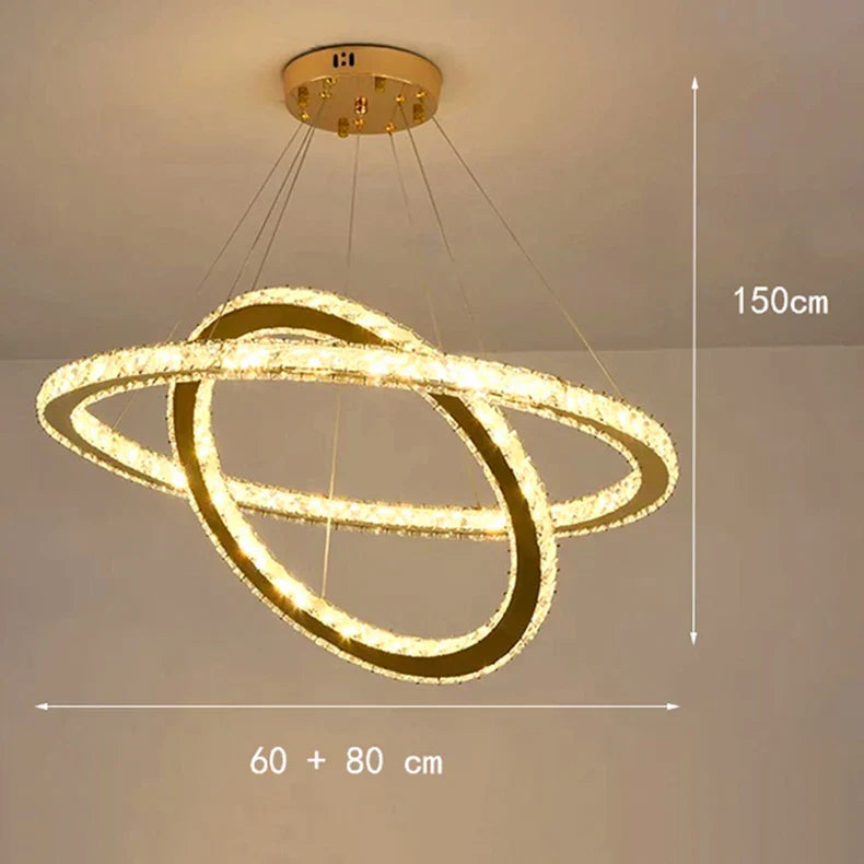 Modern LED chandelier with two intersecting circular crystal rings, gold mount, dimmable, suspended by cables.