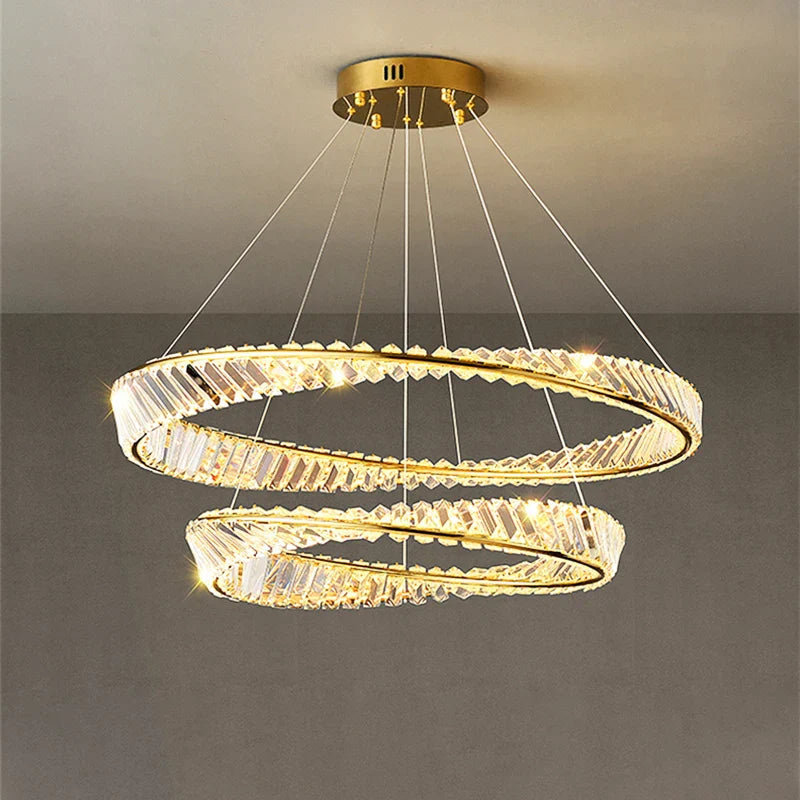 Gold crystal chandelier with two overlapping rings, suspended by wires, emits warm white light.